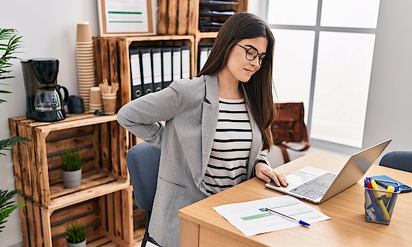 Zu sehen ist eine Frau mit Rückenschmerzen, die arbeitet.