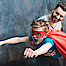 Ein Vater hält seinen Sohn hoch. Er hat einen Supermann-Umhang an und lernt fliegen, wie ein Held.