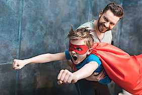 Ein Vater hält seinen Sohn hoch. Er hat einen Supermann-Umhang an und lernt fliegen, wie ein Held.