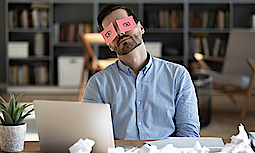 Ermüdeter Geschäftsmann sitzt am Schreibtisch mit Laptop. Er hat ein Post-it mit gemalten Augen auf dem Gesicht. 