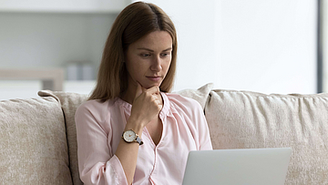 Eine Frau sitzt auf einem Sofa. Auf ihrem Schoß liegt ein aufgeklappter Laptop.