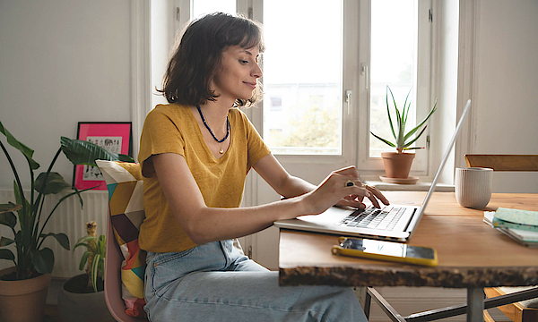 Frau ist im Homeoffice, sie sitzt am Esszimmertisch. Der geöffnete Laptop steht vor ihr.