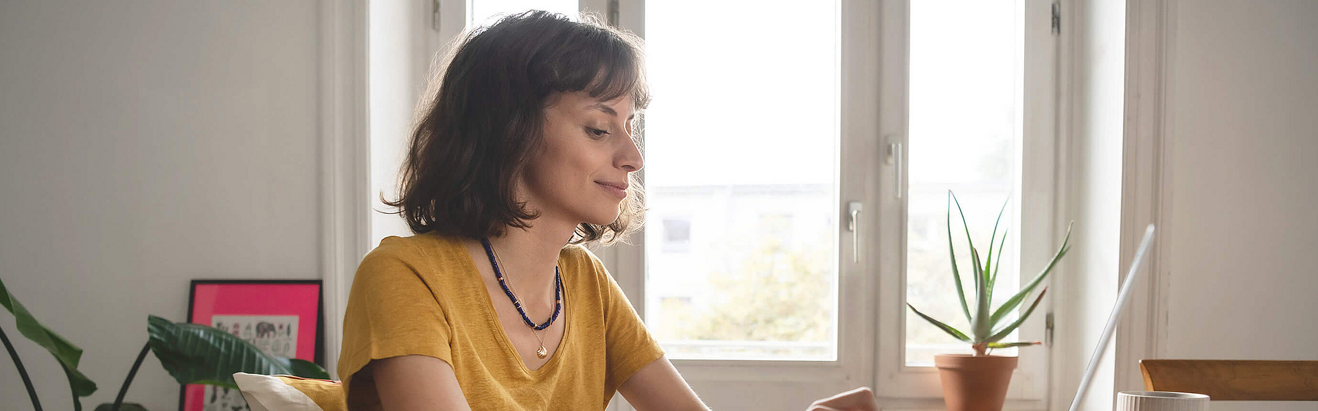 Junge Frau sitzt am Tisch mit einen geöffneten Laptop. Sie schreibt der mhplus ihre Fragen über das Kontaktformular.