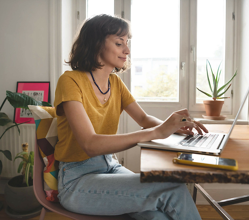 Frau ist im Homeoffice, sie sitzt am Esszimmertisch. Der geöffnete Laptop steht vor ihr.