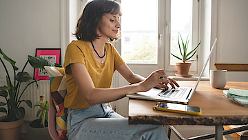 Frau ist im Homeoffice, sie sitzt am Esszimmertisch. Der geöffnete Laptop steht vor ihr.