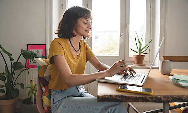 Frau ist im Homeoffice, sie sitzt am Esszimmertisch. Der geöffnete Laptop steht vor ihr.