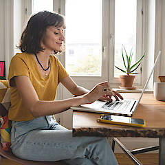 Frau ist im Homeoffice, sie sitzt am Esszimmertisch. Der geöffnete Laptop steht vor ihr.