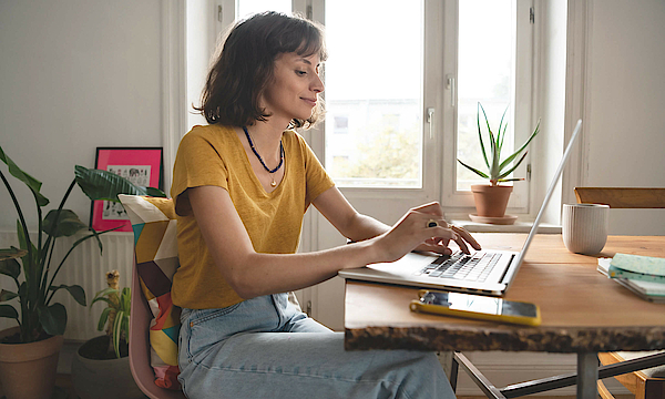 Frau ist im Homeoffice, sie sitzt am Esszimmertisch. Der geöffnete Laptop steht vor ihr.