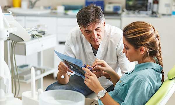 Ein Zahnarzt bespricht mit einer Patientin die anstehende Behandlung. Der Arzt hält ein Röntenbild in der Hand und zeigt darauf.