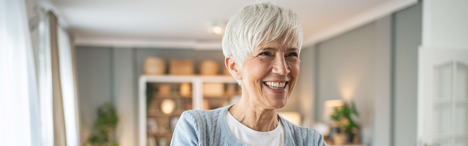 Reife freundlich lächelnde Frau steht mit gekreuzten Armen im Raum.