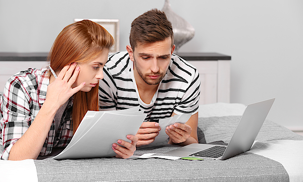 Ein Mann und eine Frau liegen auf dem Bett. Vor ihnen ist ein Laptop. Sie halten Unterlagen in Händen. Sie machen ihre Steuererklärung.