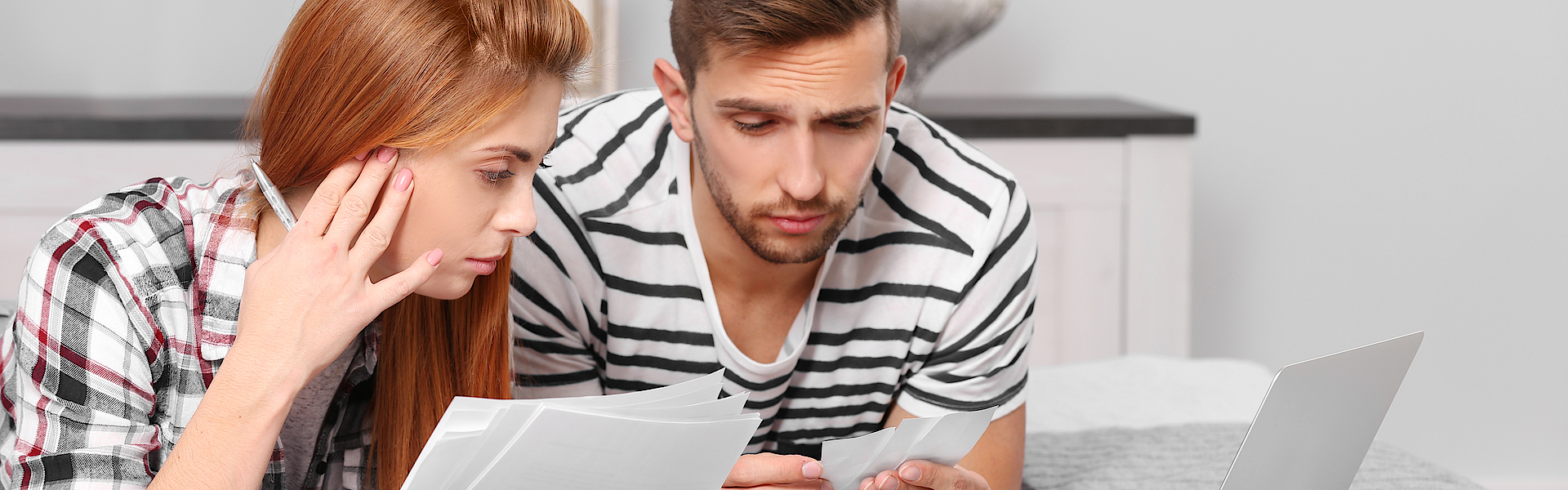 Ein Mann und eine Frau liegen auf dem Bett. Vor ihnen ist ein Laptop. Sie halten Unterlagen in Händen. Sie machen ihre Steuererklärung.