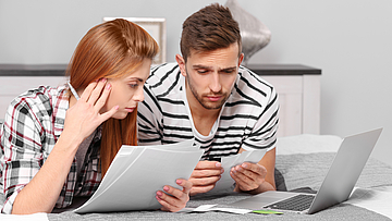 Ein Paar vor dem Laptop, sie halten Unterlagen für die Steuererklärung in Händen.