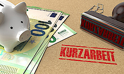 Auf einem braunen Untergrund liegen links 100€-Scheine, auf denen ein weißes Sparschwein steht. Rechts liegt ein benutzter Stempel mit "Kurzarbeit". Davor steht auf dem braunen Untergrund die Aufschrift "Kurzarbeit". Hinter dem Stempel liegen zwei Büroklammern.