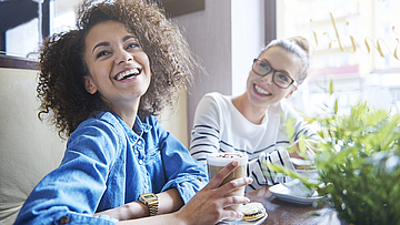 Zwei glückliche Frauen sind in einem Bistro. Die Frau im Vordergrund trinkt einen Latte Macchiato.