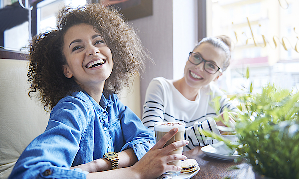 Zwei glückliche Frauen sind in einem Bistro. Die Frau im Vordergrund trinkt einen Latte Macchiato.