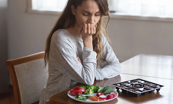 Eine Frau sitzt an einem Tisch. Vor ihr liegt eine Schachtel mit Pralinen und ein Teller mit Gemüse uns Salat. Sie muss sich entscheiden, was sie essen möchte.