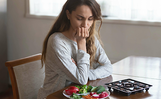 Eine Frau sitzt an einem Tisch. Vor ihr liegt eine Schachtel mit Pralinen und ein Teller mit Gemüse uns Salat. Sie muss sich entscheiden, was sie essen möchte.