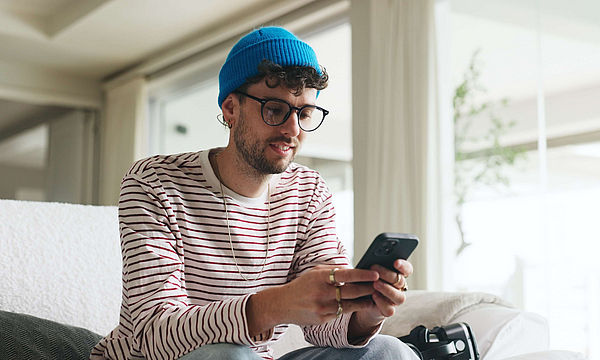 Junger Mann benützt das Smartphone er sitzt auf der Couch.