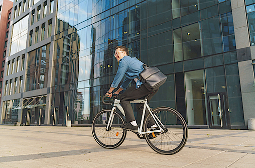 Ein lächelnder junger Mann fährt mit dem Fahrrad an einem modernen Glasgebäude vorbei.