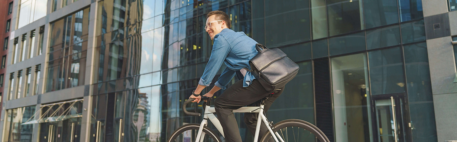 Ein lächelnder junger Mann fährt mit dem Fahrrad an einem modernen Glasgebäude vorbei.