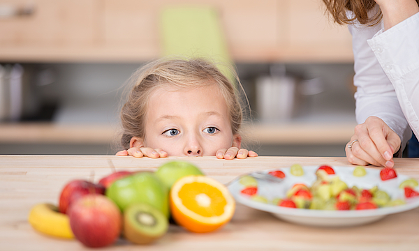 Kleines Kind etwas größer als der Küchentisch kann gerade so den Obstteller auf diesem betrachten. Am rechten Rand des Bildes sieht man Umrisse einer Frau.