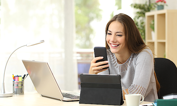 Eine junge Frau mit braunen Haaren sitzt an einem Laptop. Sie hat ein Smartphone in der Hand. Vor ihr steht ein Tablet. Die Frau arbeitet.