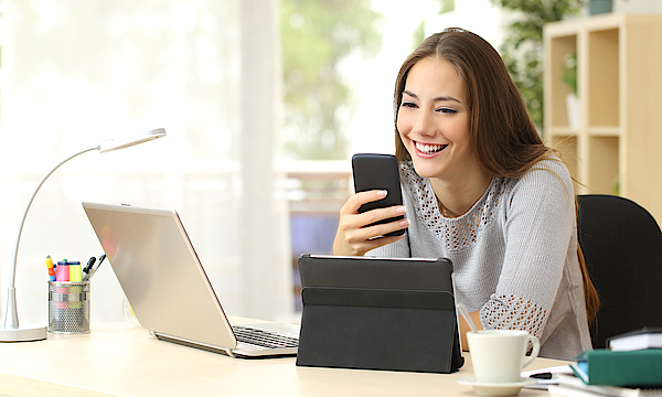 Eine junge Frau mit braunen Haaren sitzt an einem Laptop. Sie hat ein Smartphone in der Hand. Vor ihr steht ein Tablet. Die Frau arbeitet.