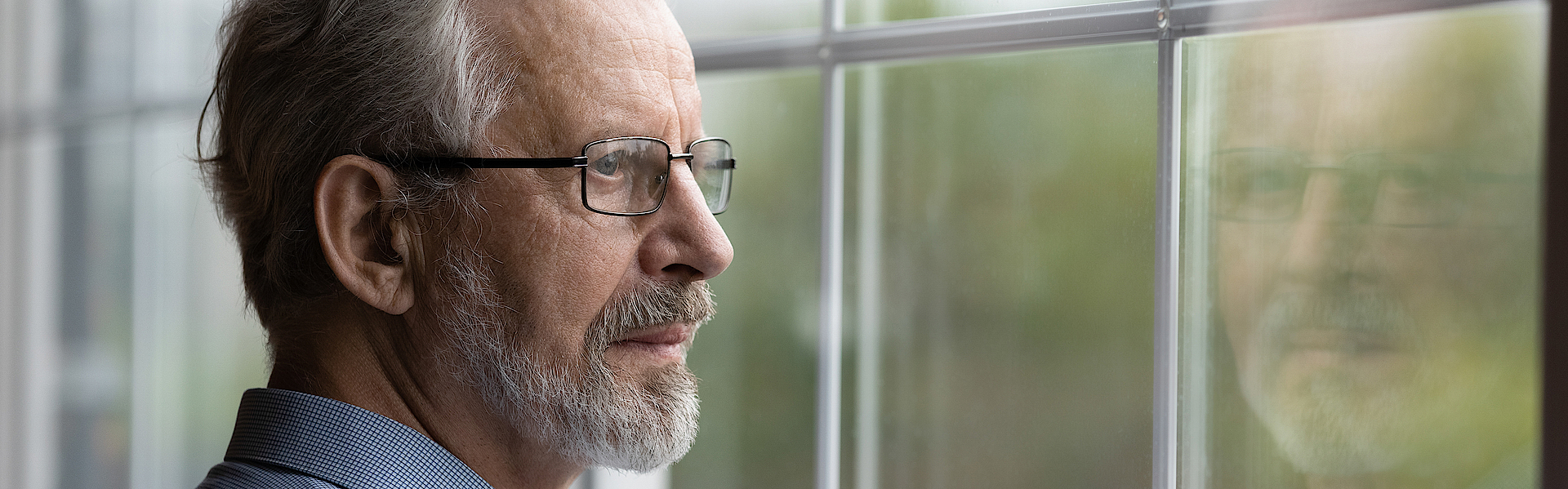 Ein älterer Mann mit grauen Haaren schaut nachdenklich aus dem Fenster. Er trägt einen Bart und eine Brille.