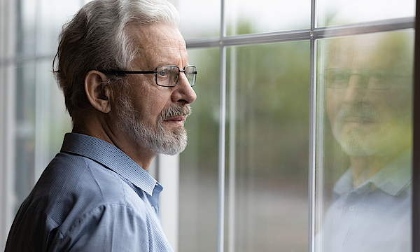 Ein älterer Mann mit grauen Haaren schaut nachdenklich aus dem Fenster. Er trägt einen Bart und eine Brille.