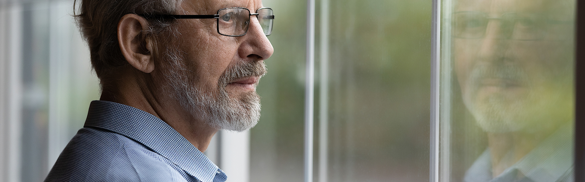 Ein älterer Mann mit grauen Haaren schaut nachdenklich aus dem Fenster. Er trägt einen Bart und eine Brille.