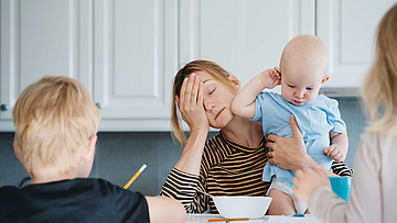 Junge erschöpfte Mutter sitzt mit zwei Kindern am Tisch und leidet an Mental Load