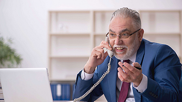 Ein Mitarbeiter in Anzug und Krawatte sitzt vor einem Laptop. Er telefoiniert aufgeregt. Sein Kopf ist rot angelaufen. Er regt sich auf.