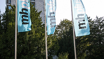 Mhplus-Hochhaus in Ludwigsburg mit drei Flaggen Foto der mhplus-Hauptzentrale in Ludwigsburg: Büro-Hochhaus in der Sonne mit Bäumen und drei Fahnen davor
