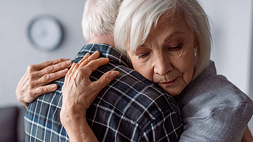 Ein älteres Paar umarmt sich. Der Mann steht mit dem Rücken zum Betrachter. Die Frau hat ihre Augen geschlossen. Ihre Hände liegen auf dem Rücken ihres Partners.