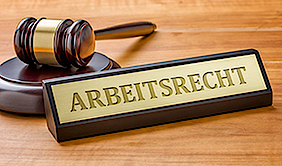 Ein Richterhammer auf einem Holztisch. Davor liegt ein schwarz-goldenes Schild mit der Gravur "Arbeitsrecht" in Großbuchstaben.