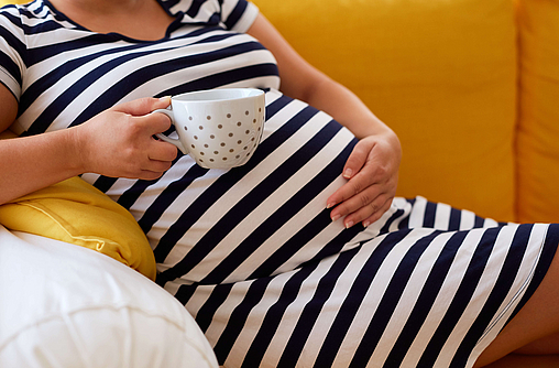 Schwangere Frau im gestreiften Kleid hält eine Tasse in der Hand.