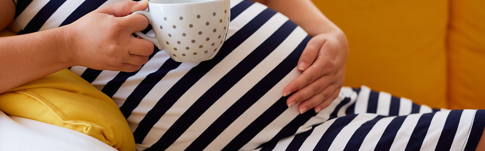 Schwangere Frau im gestreiften Kleid hält eine Tasse in der Hand.