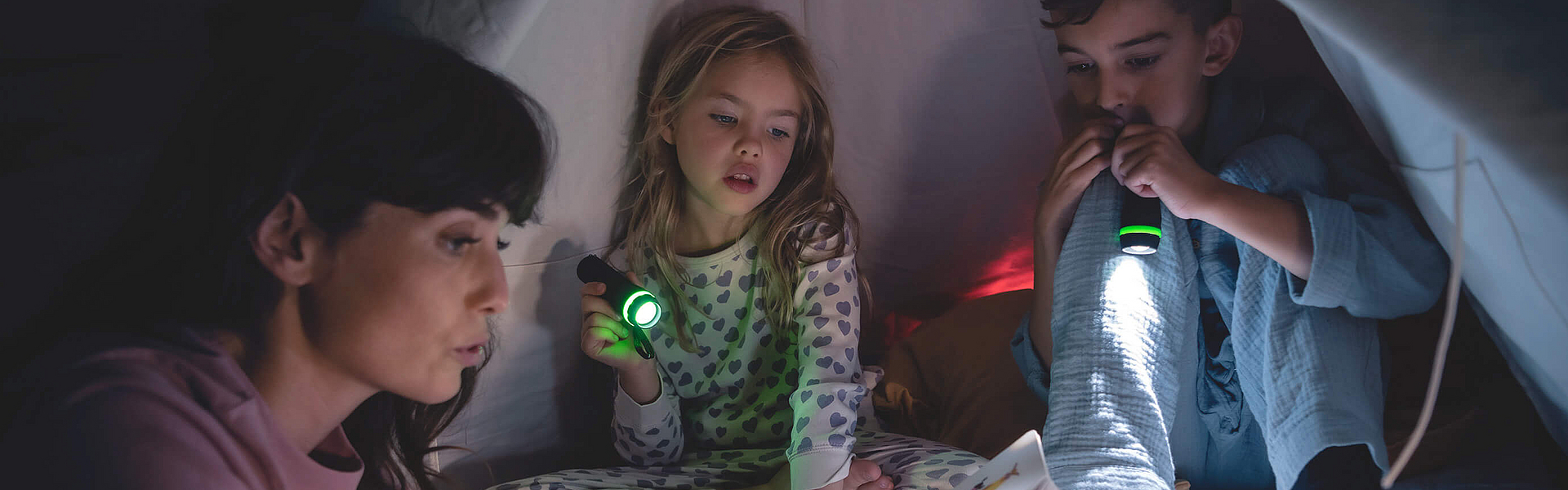 Eine Familie sitzt bei Nacht im Zelt die Kinder leuchten mit der Taschenlampe. Sie sind bei der mhplus versichert und nutzen die Vorteile für Familien.