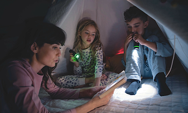 Eine Familie sitzt zusammen in einem Zelt. Es ist dunkel, der Vater und die Tochter leuchten mit einer Taschenlampe, die Mutter liest etwas aus einem Buch vor.