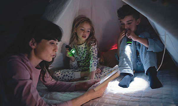 Eine Familie sitzt zusammen in einem Zelt. Es ist dunkel, der Vater und die Tochter leuchten mit einer Taschenlampe, die Mutter liest etwas aus einem Buch vor.