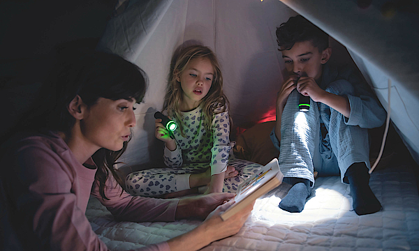 Eine Familie sitzt zusammen in einem Zelt. Es ist dunkel, der Vater und die Tochter leuchten mit einer Taschenlampe, die Mutter liest etwas aus einem Buch vor.