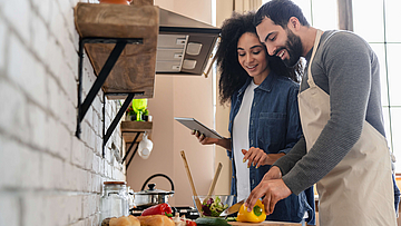 Ein Paar steht in der Küche. Er schneidet Gemüse, sie hat ein Tablet in der Hand.