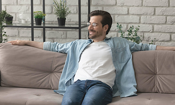 Ein Mann sitzt auf einem beigen Sofa. Er hat ein Jeanshemd und ein weißes T-Shirt an. Seine Arme liegen auf der Sofakante. Er schaut entspannt und lächelt.