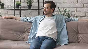 Ein Mann sitzt auf einem beigen Sofa. Er hat ein Jeanshemd und ein weißes T-Shirt an. Seine Arme liegen auf der Sofakante. Er schaut entspannt und lächelt.