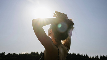 Eine junge Frau schaut von hinten mit verschränkten Armen über dem Kopf in die blendende Sonne mit einem Wald im Hintergrund. 
