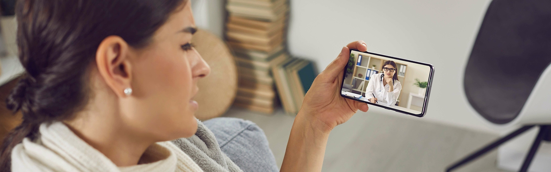 Eine Frau ist krank. Sie hält ein digitales Fiebertermometer in der rechten Hand. In der linken Hand hält sie ein Smartphone. Sie nimmt darüber an einer Videosprechstunde mit einem Arzt teil.