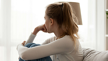 Junge Frau sitzt nachdenklich auf der Couch und sieht aus dem Fenster.