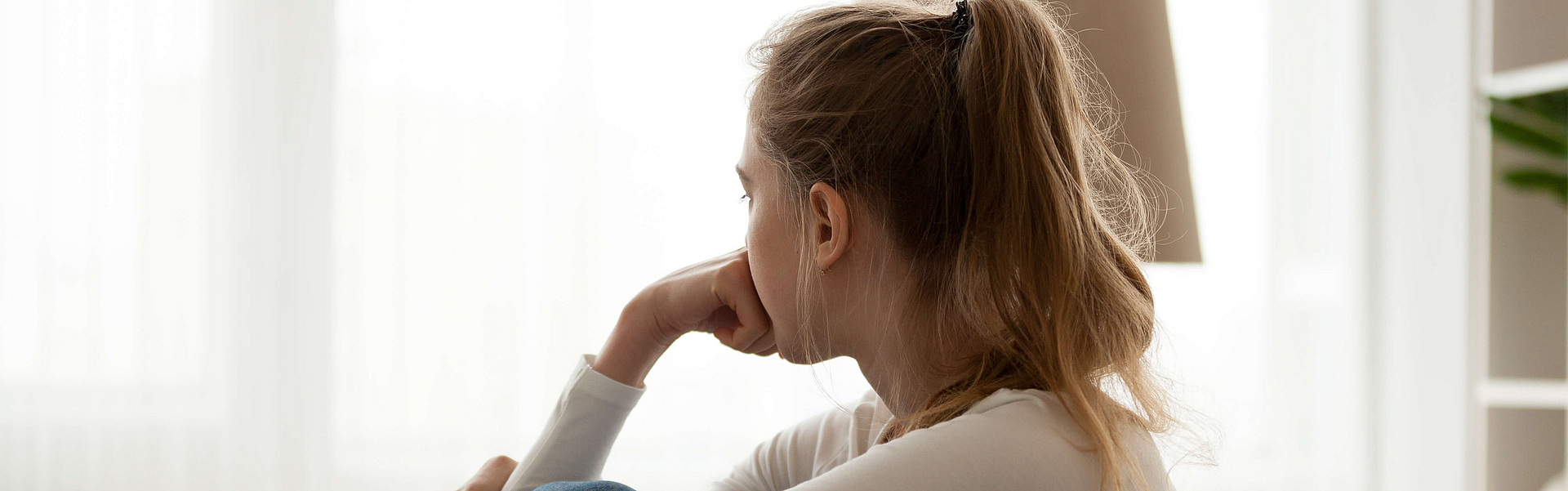 Seitliche Sicht auf eine junge Frau, die nachdenklich auf der Couch sitzt und aus dem das Fenster sieht.