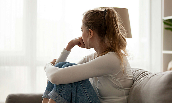 Seitliche Sicht auf eine junge Frau, die nachdenklich auf der Couch sitzt und aus dem das Fenster sieht.